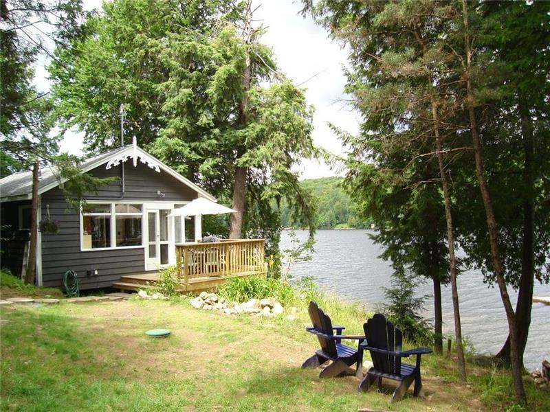 A small lakeside cottage with a wooden deck, Adirondack chairs on the lawn, and direct waterfront access.